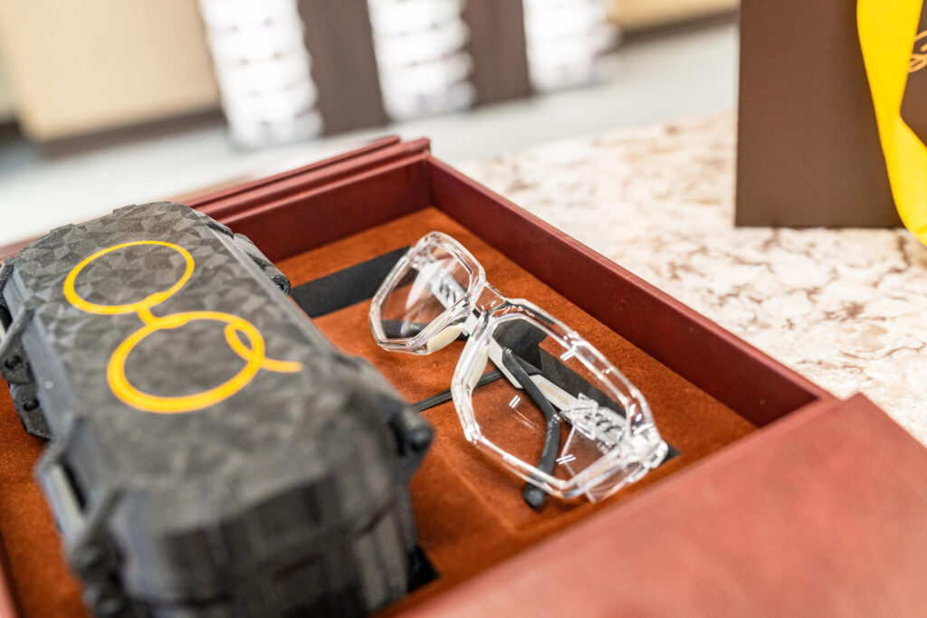 Grey felt glasses case with a gold logo, paired with clear eyeglass frames positioned behind it on a clean, minimal surface