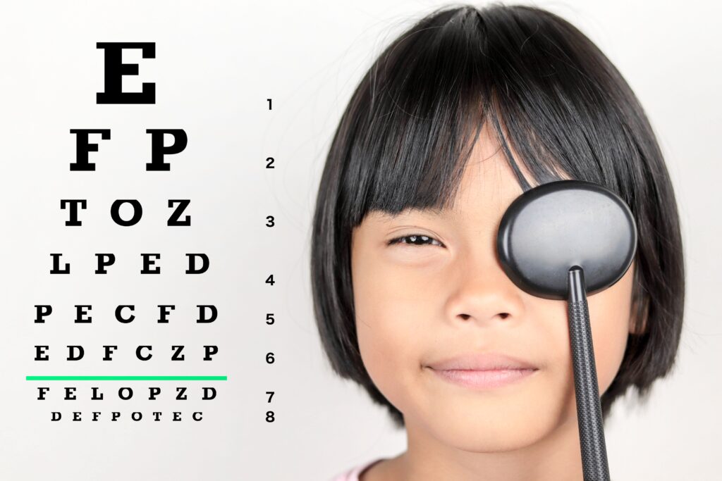 9-10-year-old Asian girl with a bob haircut covering one eye for an eye test, with an eye chart visible over her right shoulder.